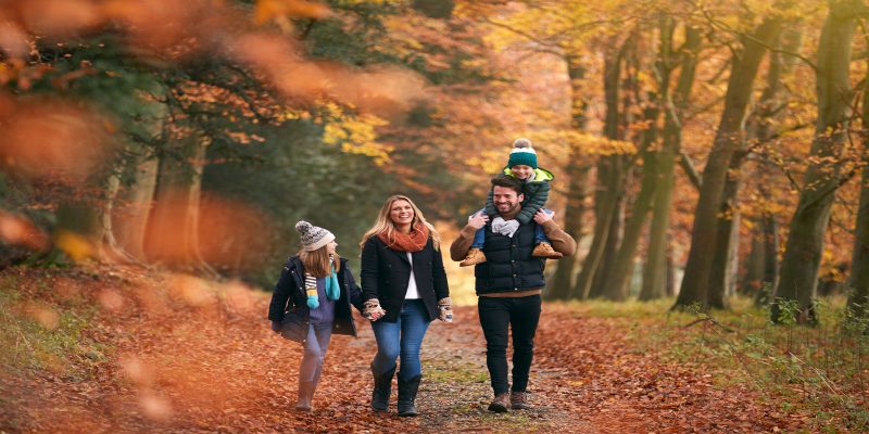 Una familia sonriente paseando al aire libre en un día de otoño, rodeada de árboles y hojas caídas en el suelo.