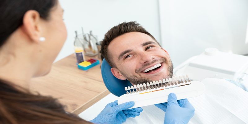 Hombre joven sentado en un consultorio mientras una odontóloga revisa la escala de colores para realizarle un blanqueamiento dental.