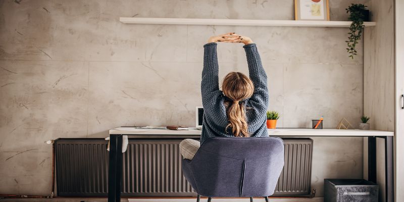 Mujer joven estirando la espalda mientras está sentada frente a su computadora, tomando una pausa durante el trabajo.