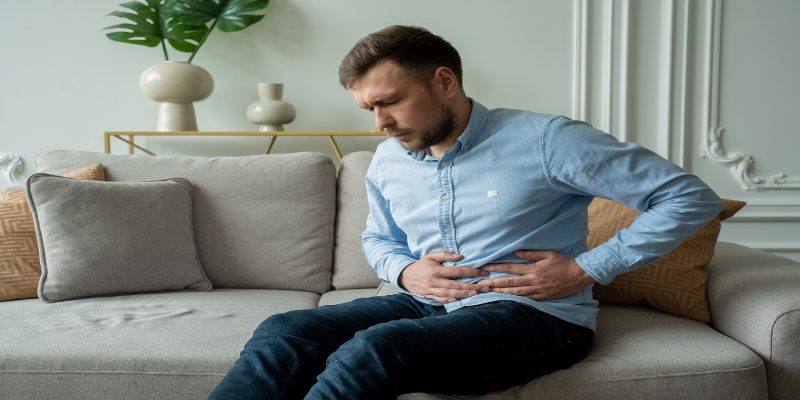 Hombre sentado en un sillón, tomándose el estómago y mostrando signos de dolor.